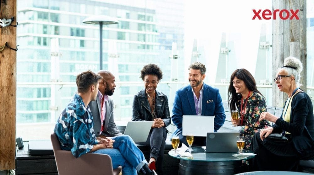 Un groupe de sept personnes assises ensemble, conversant et travaillant sur des ordinateurs portables avec une ville en arrière-plan Un groupe de sept personnes assises ensemble, conversant et travaillant sur des ordinateurs portables avec une ville en arrière-plan