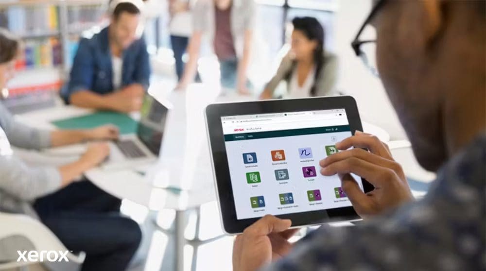 Plateforme Xerox® Workflow Central : accès mobile intuitif Une personne tient une tablette affichant l'interface Xerox Workflow Central avec diverses icônes d'application, tandis que d'autres personnes travaillent dans l'arrière-plan flou, mettant en évidence l'accès mobile aux solutions de flux de travail.